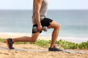 Lunge leg workout with dumbbells weights. Fitness man doing lunges training legs on beach summer outdoors with weight.