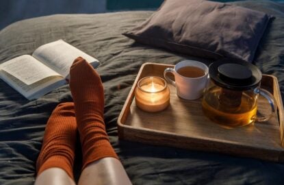 Soft photo of woman`s legs in the woolen socks on the bed with book and cup of tea and candle on the tray. Interior and home cosiness concept. Top view